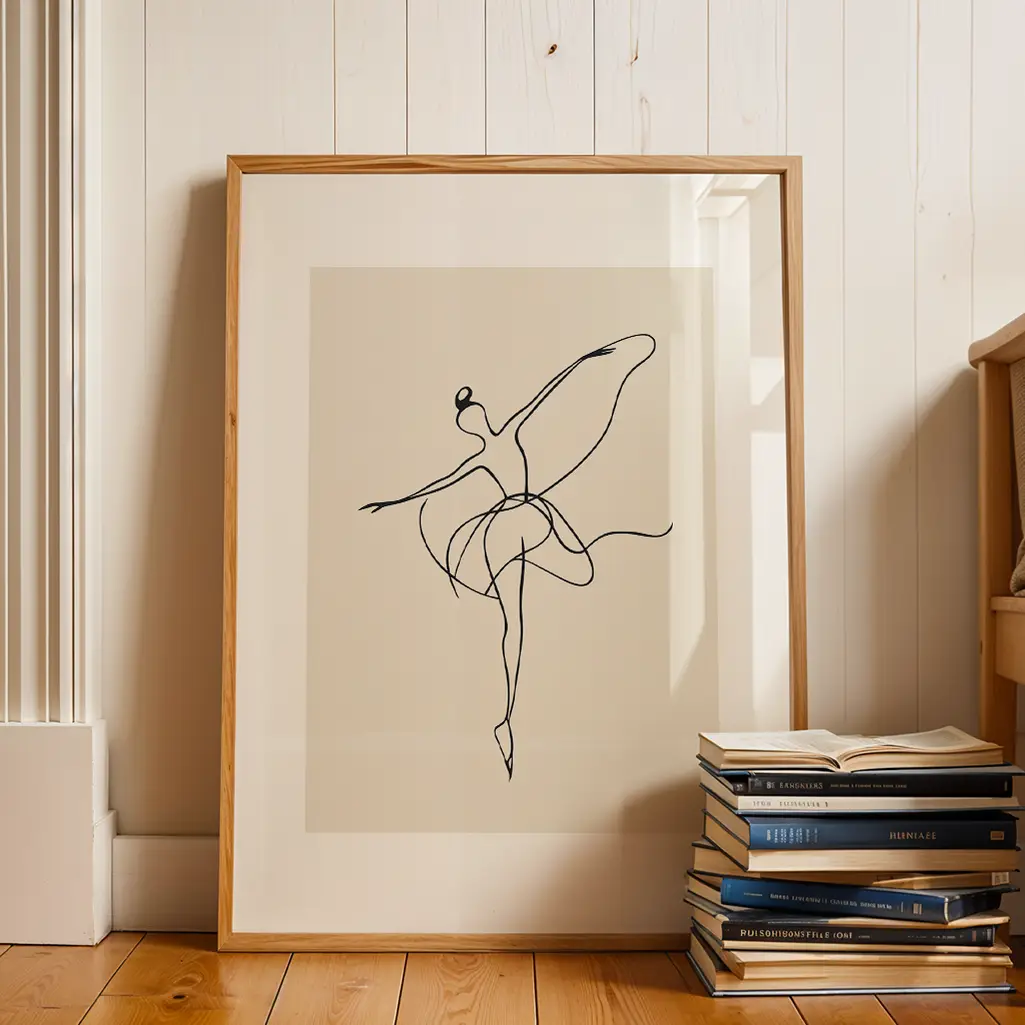 Framed ballerina line art print on a wooden floor next to a stack of books, against a white panel wall.